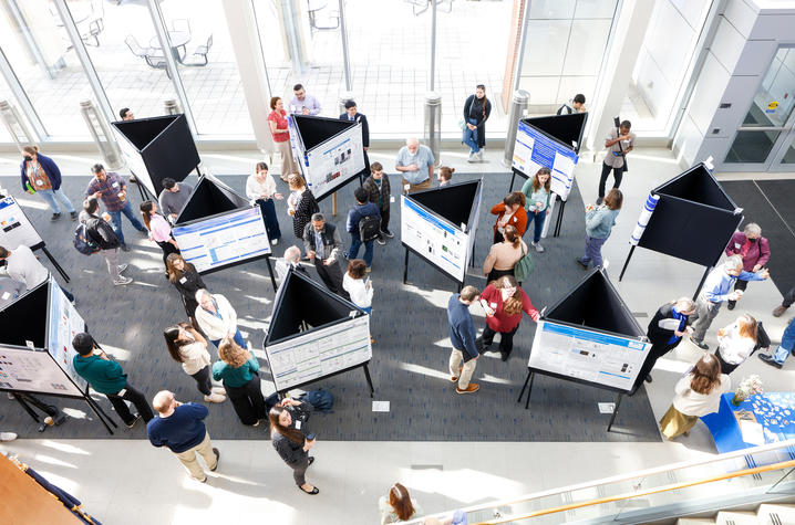 Overhead view of a busy indoor research poster session with numerous triangular poster boards arranged in rows. Attendees stand in small groups discussing the displayed research in a bright, sunlit atrium.
