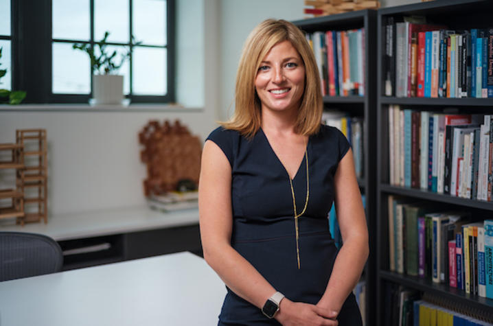 Person standing in a modern office, wearing a dark dress, with bookshelves in the background.