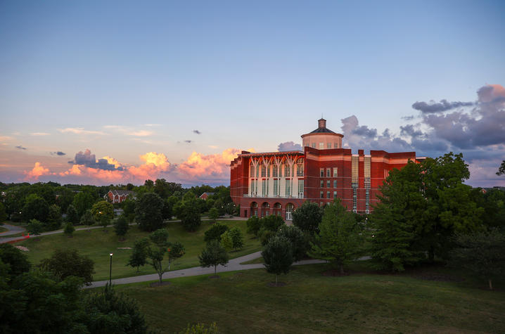 Red-brick campus building seen at sunset with trees and open green space in the foreground and colorful clouds in the sky.
