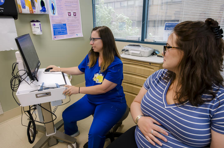 Image of UK provider measuring a pregnant patient’s belly. A health care professional in blue scrubs uses a computer beside a seated pregnant patient in an exam room.