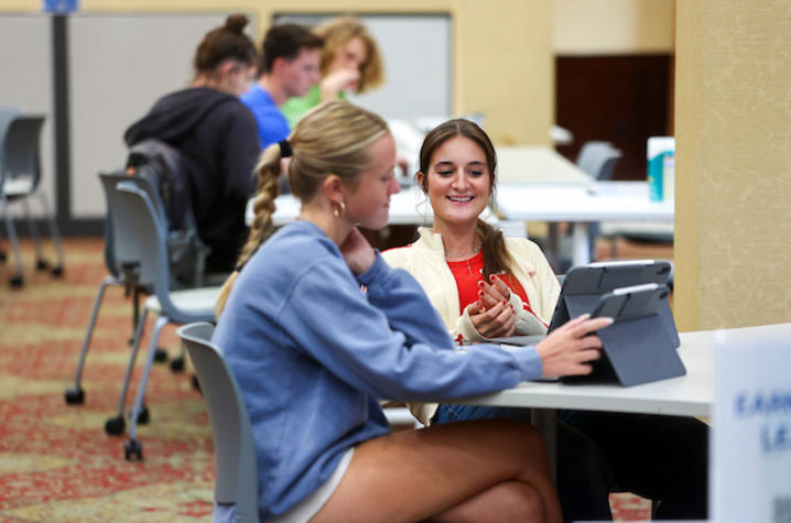 Two students sit at a table, reviewing content on a tablet together while others work in the background.