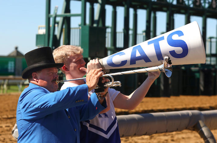 Two people at a horse racetrack, one playing a trumpet and one shouting into a megaphone labeled “CATS.”