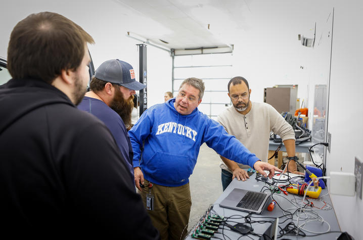 Group of people gathered around a workbench, looking at electronics and tools in a workshop.