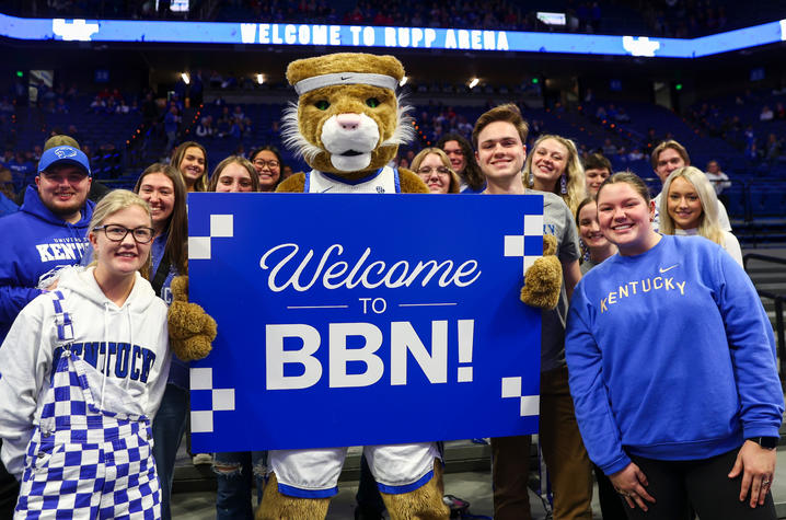A group of students standing with the Kentucky Wildcat mascot. The mascot is holding a blue sign that says “Welcome to BBN”