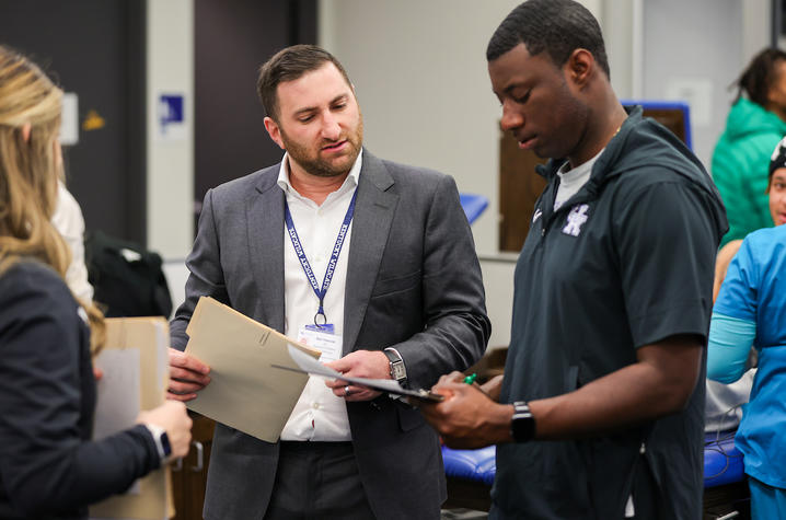 Photo of Marc Paranzino and UK Sports Cardiology team members Two people stand indoors discussing documents, one holding a folder and the other a clipboard, with others in the background.