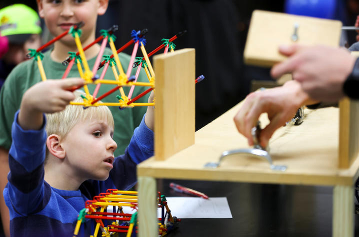 A child builds a small bridge-like structure using colorful plastic construction pieces, while an adult nearby demonstrates a wooden apparatus with metal components. Other children stand close, observing the activity.