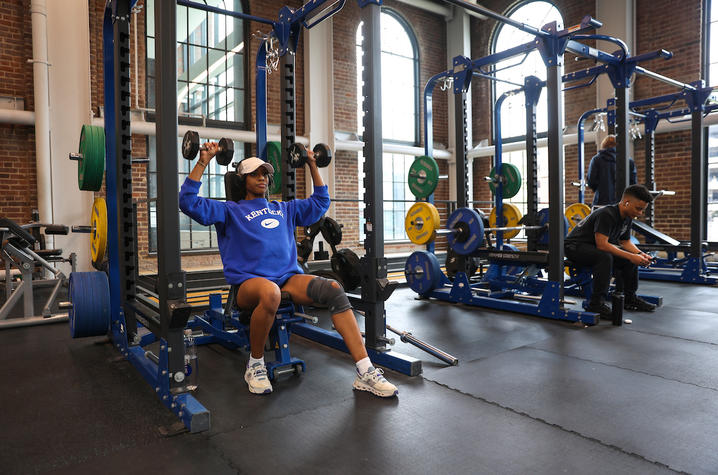 A student wearing a blue Kentucky sweatshirt lifts dumbbells while seated at a weight station, with other students and strength training equipment visible in the background.