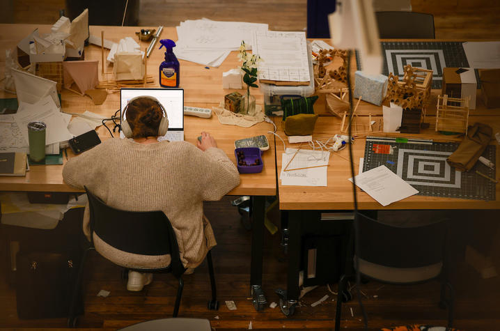 High-angle shot of a student working on a laptop at a wooden table with drafting tools and papers.
