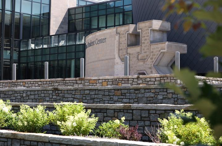 Exterior view of a modern campus building with large glass windows and stone walls, featuring a tiered stone landscape with green shrubs in the foreground and architectural details of the student center visible behind.