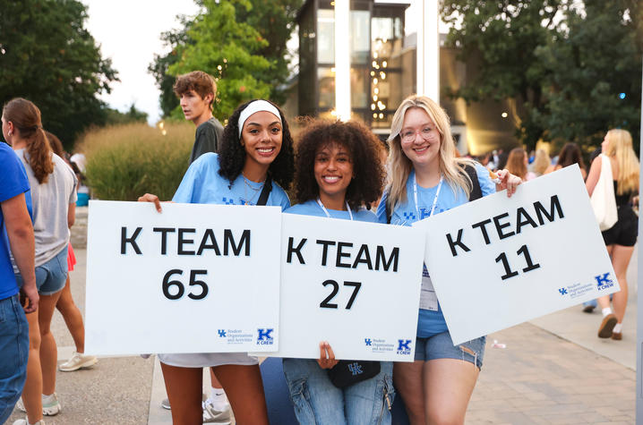 Three people hold signs reading “K TEAM 65,” “K TEAM 27” and “K TEAM 11” while standing outdoors on campus during a university event, with other attendees visible in the background.
