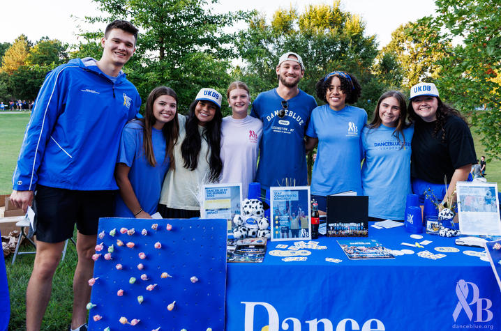 Photo of DanceBlue table at K Week Seven people in blue shirts smile behind a DanceBlue information table set up outdoors, with brochures and display boards arranged on top.