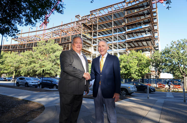 Two people stand on a sidewalk in front of a large construction site featuring a multistory steel framework of a building under development. 