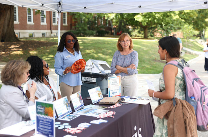 People speak with visitors at an outdoor information table under a tent on campus, with brochures, handouts and a landfill waste display.