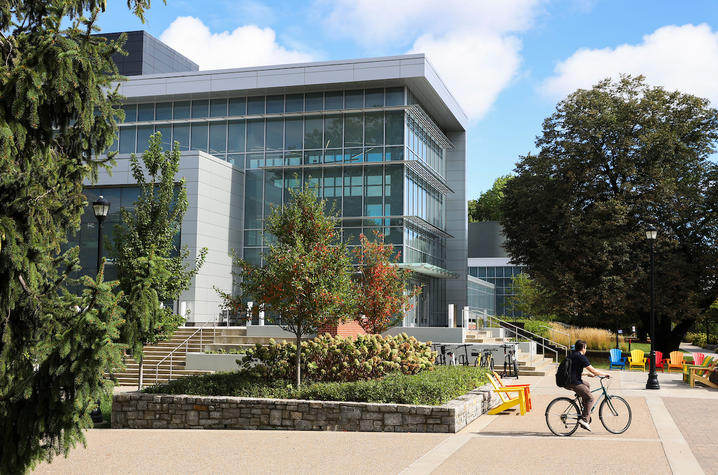 Modern glass-front academic building with large windows and a white exterior, surrounded by trees and landscaping, as a cyclist rides past on a campus sidewalk.