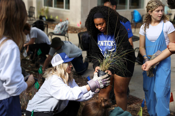 A Black woman standing and a white woman crouching, preparing to put a plant into a garden.