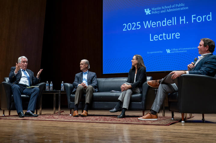2025 Ford Lecture participants Four panelists sit on a stage during a public lecture and discussion at the University of Kentucky. One speaker gestures while talking from an armchair, while the other panelists listen from a sofa and adjacent chairs. A large screen behind them.