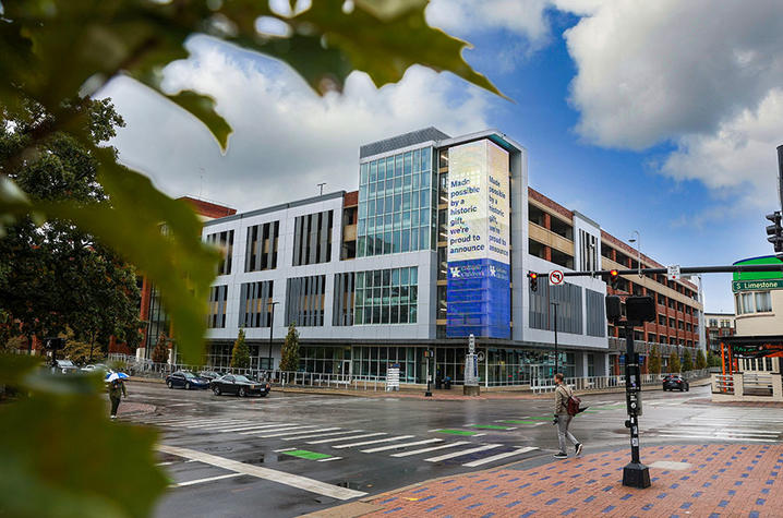 Modern multi-story academic building on a rain-slicked city corner, framed by green leaves, with pedestrians and light traffic at the intersection.