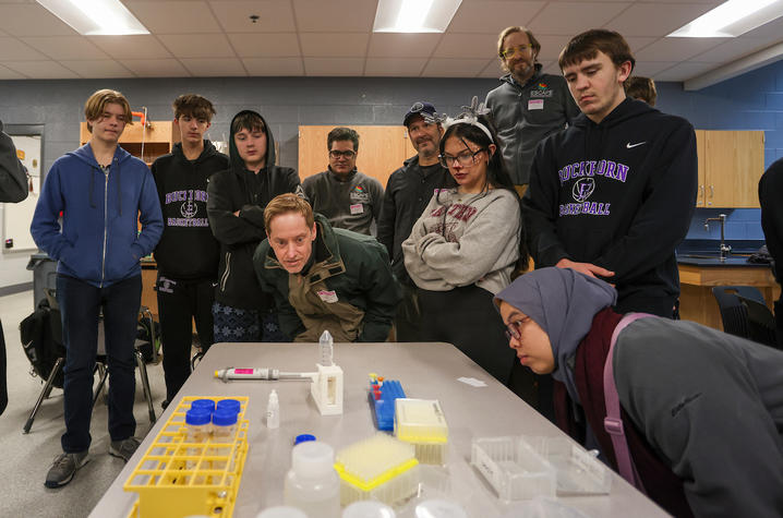 Group of students gathered around a table observing lab equipment and materials in a classroom setting.