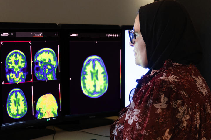 A woman wearing glasses and a headscarf studies colorful brain scan images displayed on two computer monitors in a medical research setting.