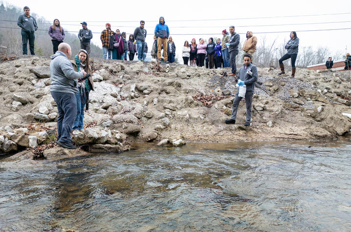 Group of people standing on a rocky bank near a stream, with two individuals in the foreground engaged in discussion or activity by the water.