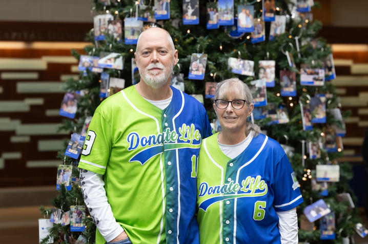 Two people wearing green and blue ‘Donate Life’ baseball jerseys standing in front of a decorated Christmas tree with photo ornaments