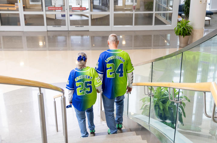 Two people walking up a staircase inside a building, wearing bright green and blue jerseys with the words ‘RECIPIENT 25’ and ‘SAVING DONOR 24’ on the back