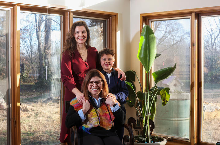 Three people indoors near large windows with sunlight, one seated and two standing, next to a tall green potted plant.