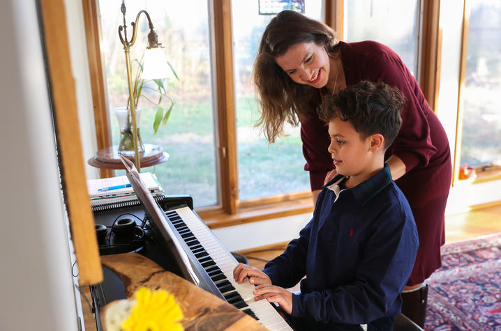 Person standing beside a child playing a piano in a sunlit room with large windows and a patterned rug.