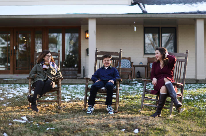 Three people sitting on wooden rocking chairs in a grassy yard with light snow, in front of a house with a covered porch.