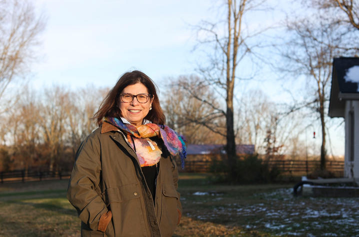 Person standing outdoors in a grassy yard wearing a dark coat and colorful scarf, with bare trees and a house in the background.
