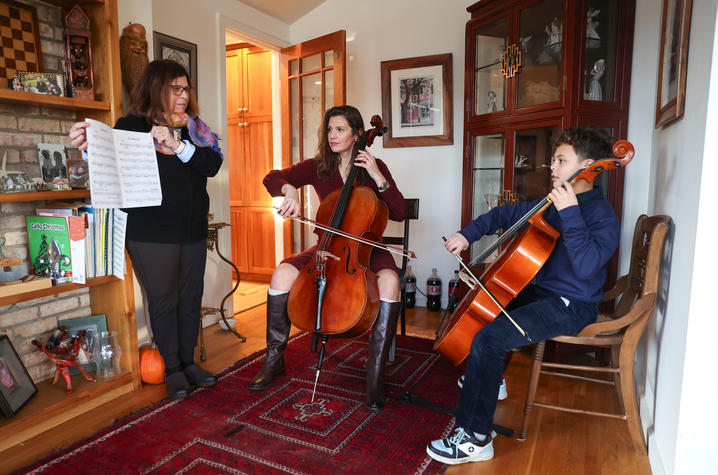 Two people playing cellos while another person holds sheet music in a cozy room with wooden floors, shelves, and a red rug.
