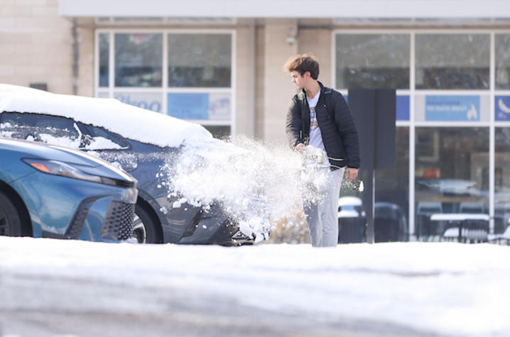 Person clearing snow from a car in a snowy parking lot near a building with large windows.