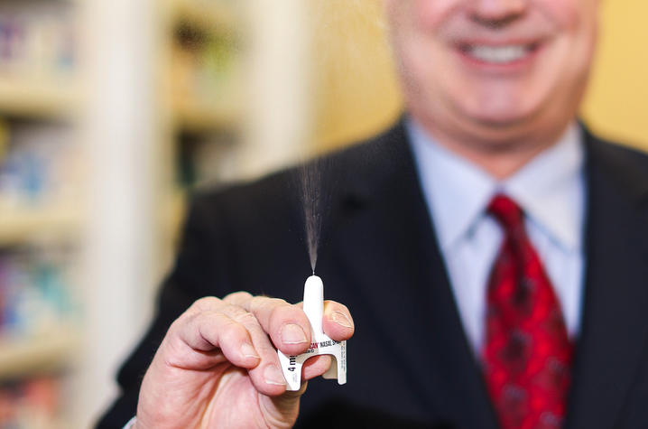 A man in a suit holds up a naloxone nasal spray device as it releases a fine mist, demonstrating how it works. Shelves of medication are softly blurred in the background, keeping focus on the overdose-reversal spray in his hand.