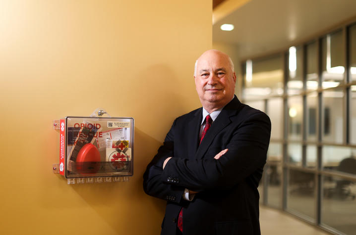 A man in a dark suit and red tie stands with his arms crossed beside a wall-mounted opioid rescue box in a brightly lit hallway. The box contains naloxone and overdose response information, with glass-walled offices visible in the background.