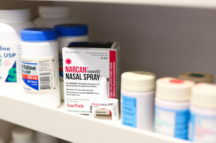 Boxes of Narcan (naloxone) nasal spray sit on a shelf alongside several prescription medication bottles. The close-up highlights the overdose-reversal medication stored among routine clinical supplies.