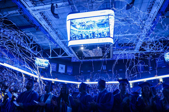Graduates in caps and gowns stand inside Rupp Arena as blue streamers fall from the ceiling, with a large scoreboard displaying the celebration overhead.