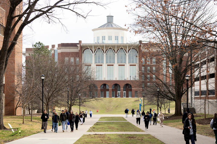 A wide campus walkway lined with leafless trees and grass leads toward a large academic building with arched windows and a central cupola, while groups of students walk along the path on an overcast day.