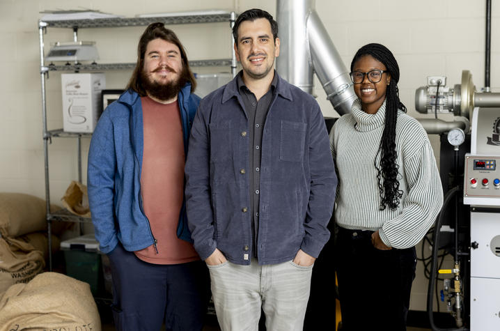 photo of faculty in the UK Coffee Lab at the UK Horticulture Research Farm Three people stand together in an industrial workspace with coffee equipment and large bags in the background. One person on the right is smiling, and all three are posed in front of machinery and shelving.