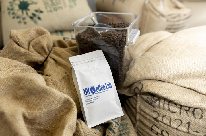 photo of UK Coffee Lab’s beans A sealed bag labeled “UK Coffee Lab” sits on top of stacked burlap sacks of coffee beans, with a clear container filled with roasted beans placed behind it.