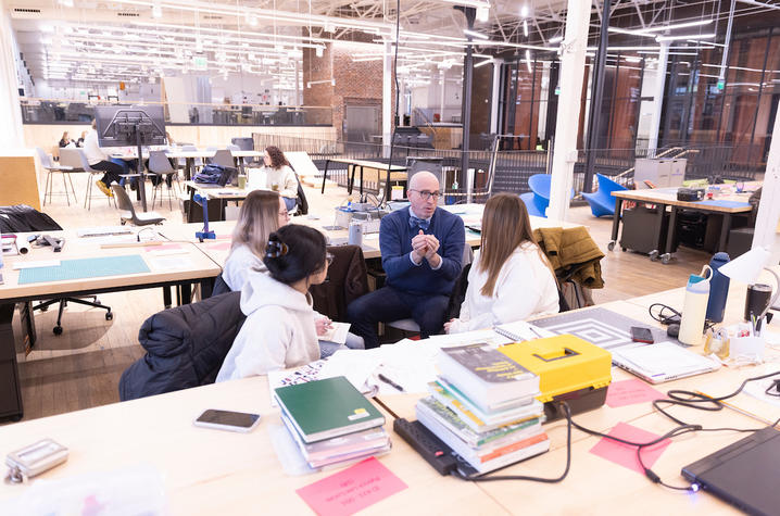 A man speaks with three students around a table covered with books, notebooks and design materials in an open workspace.