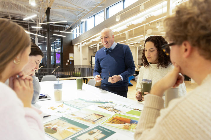A person and four students review design boards spread across a table in an open workspace.