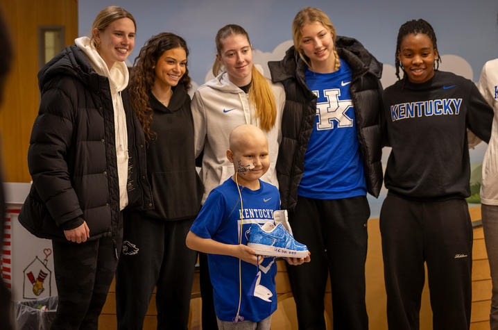 A group of people stand together indoors, posing for a photo. They are wearing casual and athletic clothing, including University of Kentucky apparel. A child at the front of the group holds up a brightly colored custom sneaker. 