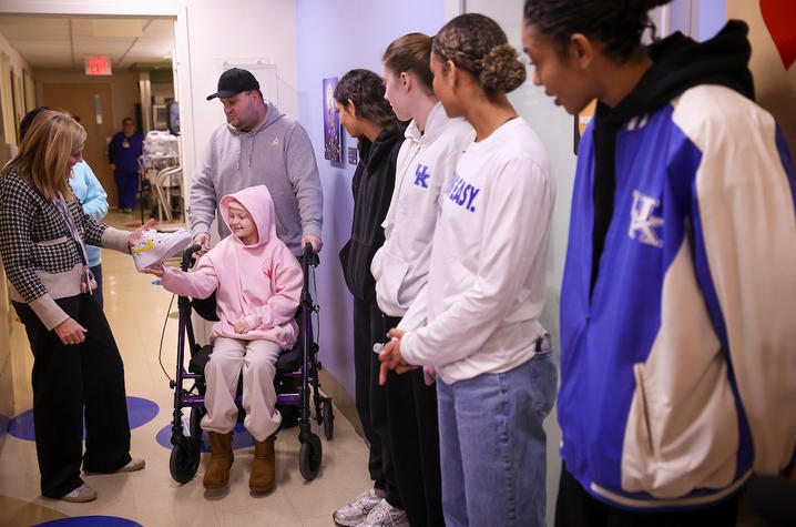 A child seated in a wheelchair reaches out to greet a group of people standing in a hallway. The child wears a pink hoodie and winter boots, and an adult stands nearby. Several individuals wearing University of Kentucky athletic apparel stand in a line.