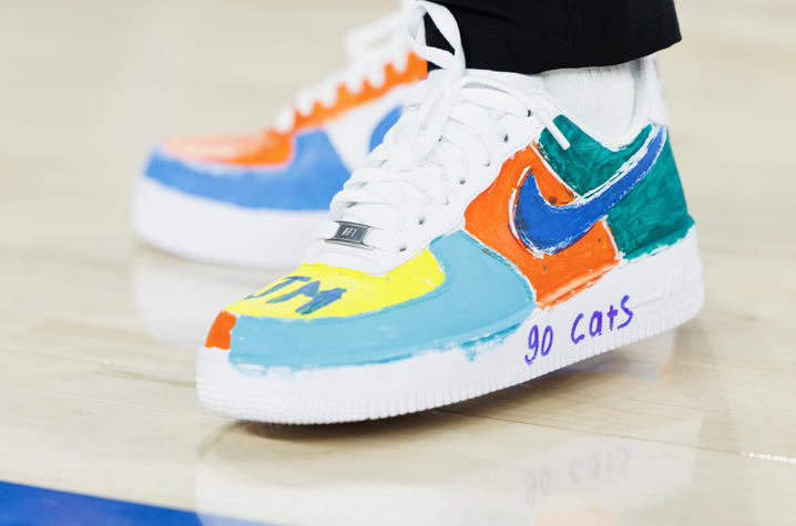 A close-up of a pair of custom-painted athletic shoes featuring bold, colorful sections in blue, orange, teal, green and yellow. One shoe includes a blue Nike swoosh and the handwritten words “go cats” on the white midsole.