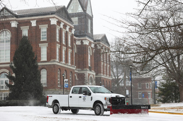 Snowplow truck clearing snow on a campus road in front of a historic brick building.