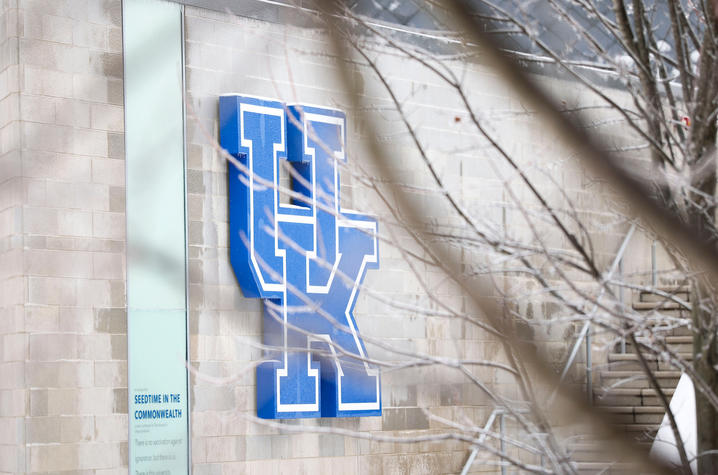 Large blue “UK” logo mounted on the exterior wall of a beige stone building, partially obscured by out‑of‑focus tree branches in the foreground.