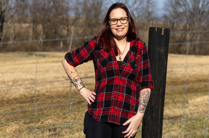 A woman stands outdoors in a grassy field, leaning slightly against a wooden post. They are wearing a red‑and‑black plaid shirt. Trees and a wire fence are in the background