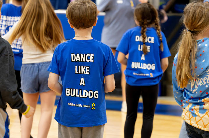 Children wearing matching blue T-shirts that read “Dance Like a Bulldog” stand in a gymnasium, facing away from the camera as they participate in a group activity.