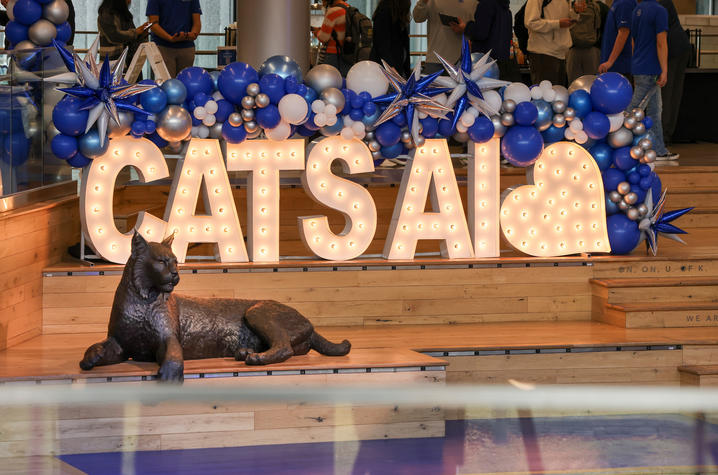 A Wildcat statue rests beside a fountain in front of a large illuminated “CATS AI” sign decorated with blue balloons at a campus event.