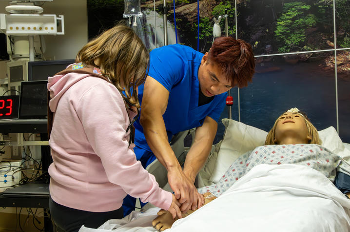A Girl Scout practices a first aid technique with guidance from a nursing instructor beside a hospital bed using a training mannequin.